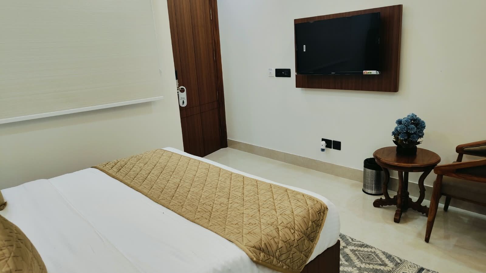 A hotel room with a bed, a television mounted on the wall, a small table with a vase of flowers, and a chair.