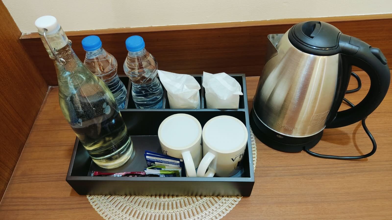 Mesita de noche en una habitación de hotel con una botella de agua, dos botellas adicionales, una caja de pañuelos, una taza, un hervidor y una pequeña bandeja con una servilleta.