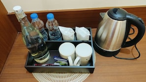 Mesita de noche en una habitación de hotel con una botella de agua, dos botellas adicionales, una caja de pañuelos, una taza, un hervidor y una pequeña bandeja con una servilleta.