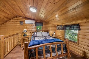 Ein Schlafzimmer in einer Blockhütte mit einem Bett, einem Nachttisch, einer Lampe und einem Fenster mit Blick auf Grünflächen.