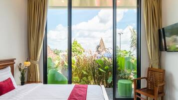 A hotel room with a large bed, a wooden chair, a TV on the wall, and a view of greenery and a building outside.