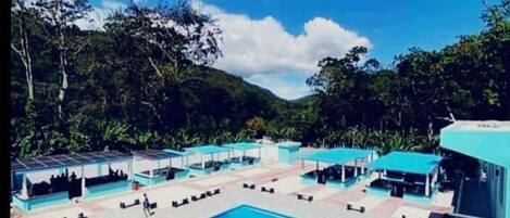 Uma área de piscina com várias seções e zonas de estar cobertas, cercada por vegetação.