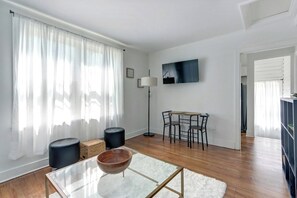 A modern living room with a glass coffee table, a black ottoman, a wooden floor, a dining area with chairs, and a flat-screen TV mounted on the wall. - Fresh Modern Apartment by Daffin (Savannah)