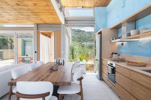 A modern kitchen with a wooden table and white chairs, a built-in oven, and a view of greenery outside. - Anassa House with Rooftop Jacuzzi (Gavalochori)