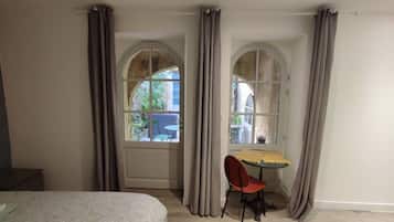 A room with a bed, a table with a chair, and two windows with curtains.