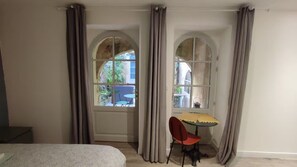 A room with a bed, a table with a chair, and two windows with curtains. - Studio on patio in Chartrons (Bordeaux)
