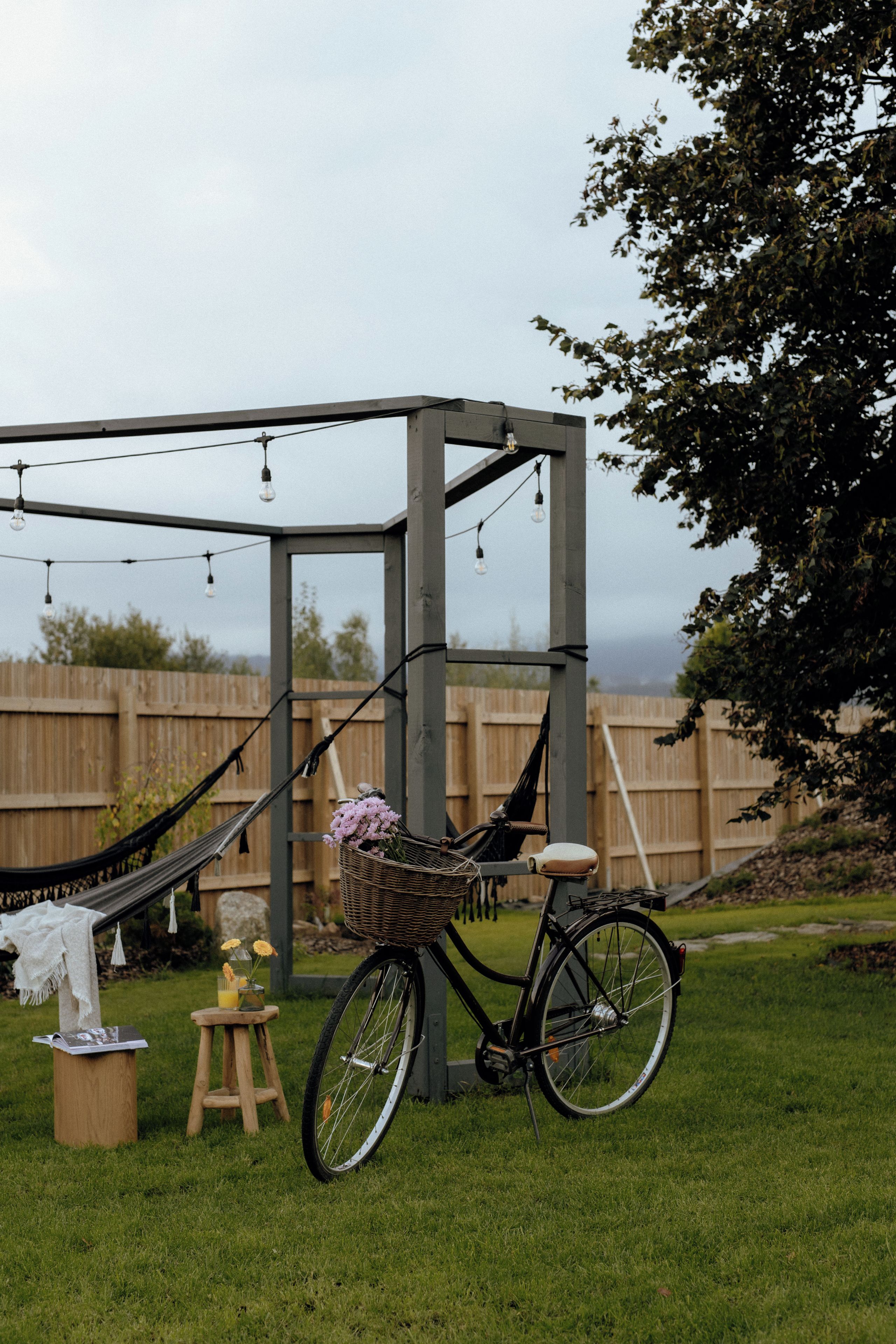 A bicycle with a basket and a hammock in a grassy outdoor area with a wooden fence in the background.