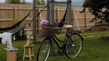 A bicycle with a basket and a hammock in a grassy outdoor area with a wooden fence in the background.