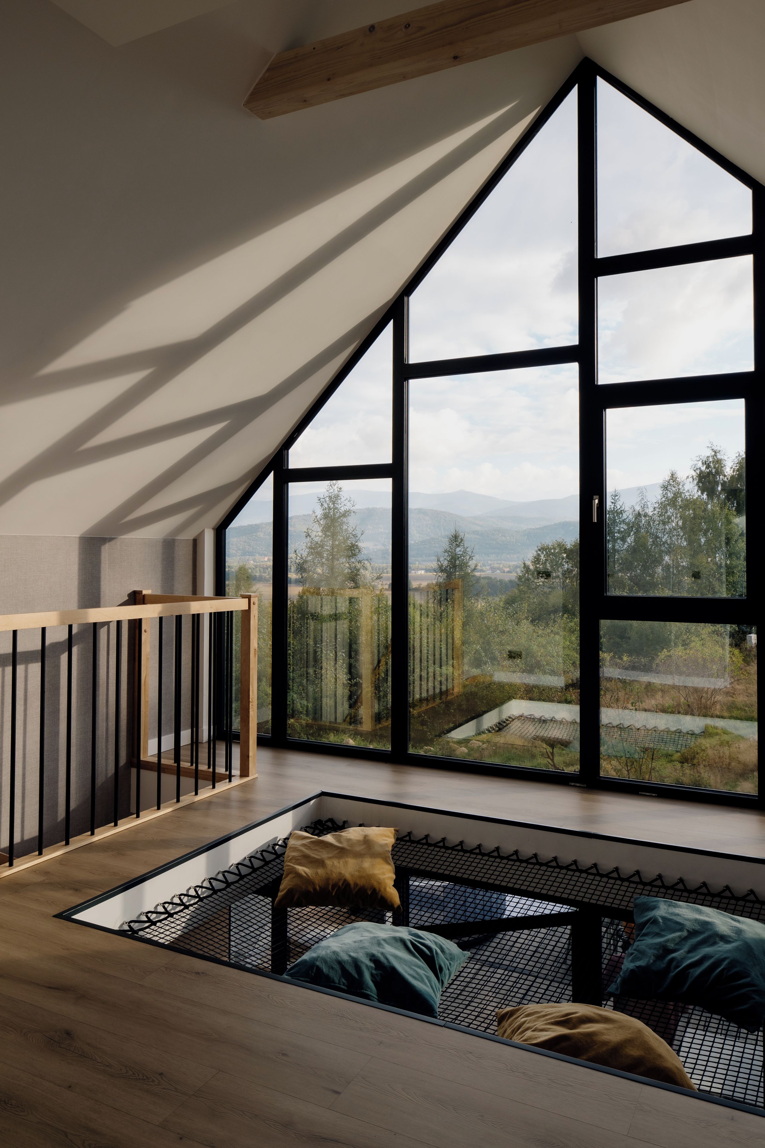 A room with a sloped ceiling, a large window with a view of trees and mountains, a wooden bench with cushions, and a metal railing.