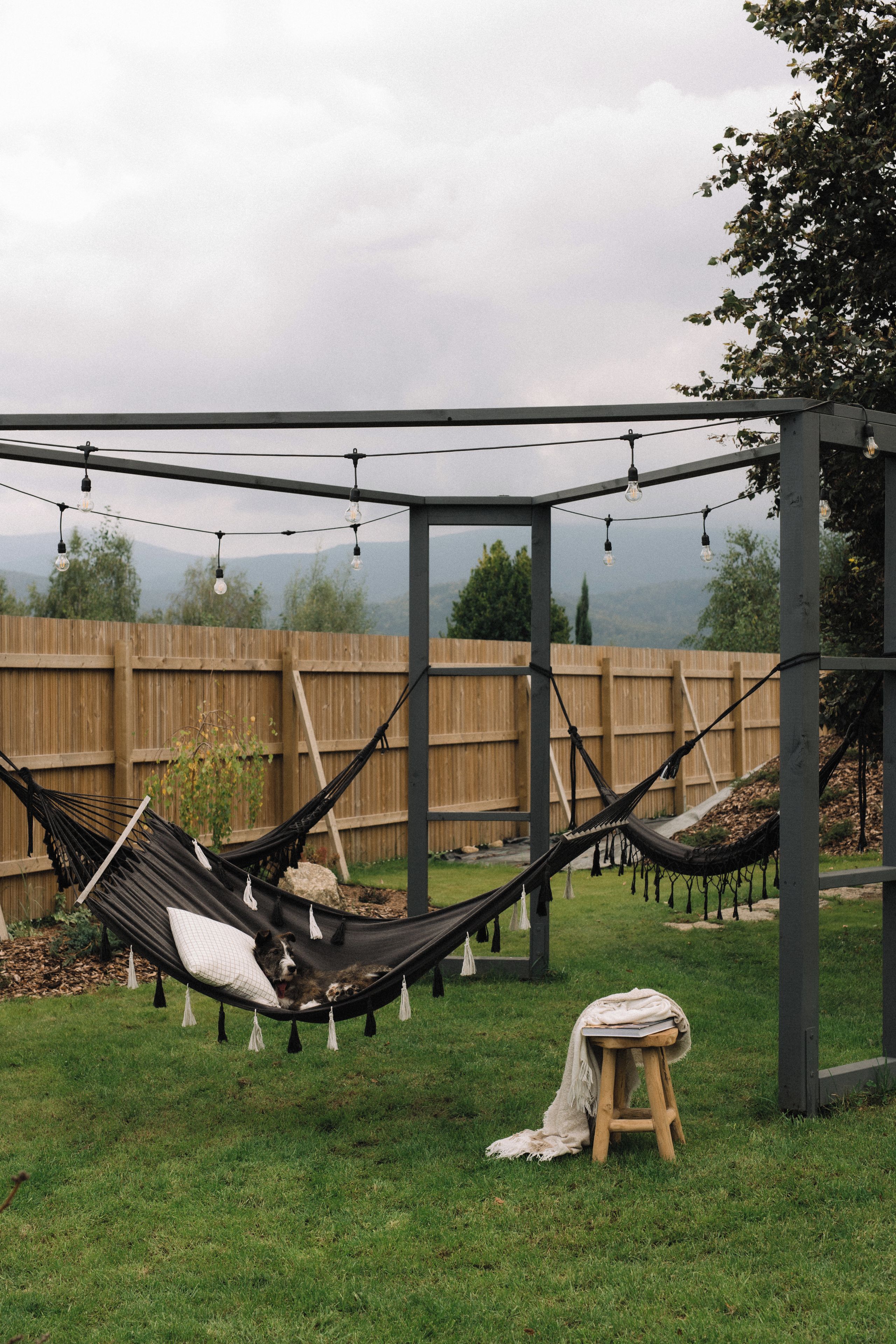 Uma rede sob uma pérgola decorada com luzes, uma cerca de madeira e um gramado.