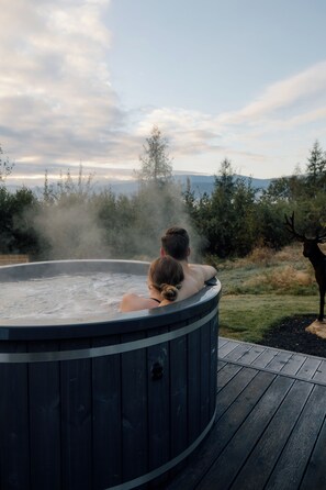 A hot tub with two people sitting inside, surrounded by a deck and a natural landscape with trees and a deer in the background.