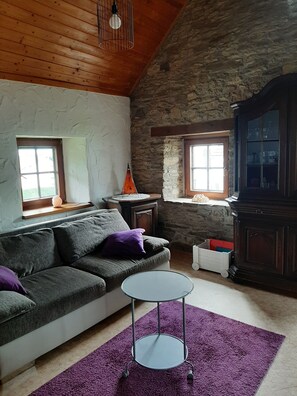 A cozy living room with a stone wall, wooden ceiling, a grey sofa with purple cushions, a small round table, and a cabinet with glass doors.