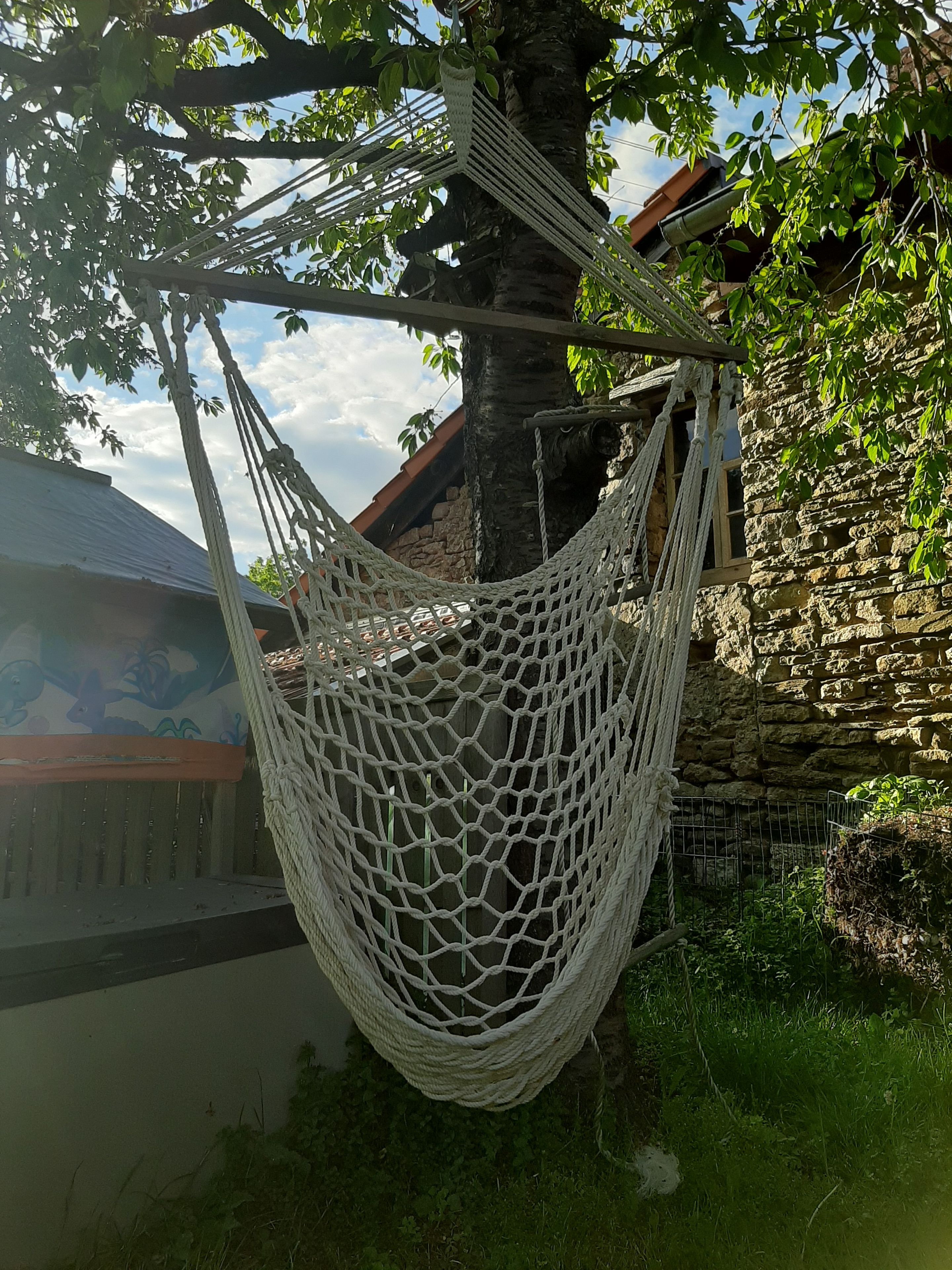 Eine Hängematte zwischen Bäumen in einem Garten mit einer Steinmauer und einem Gebäude mit Schrägdach.