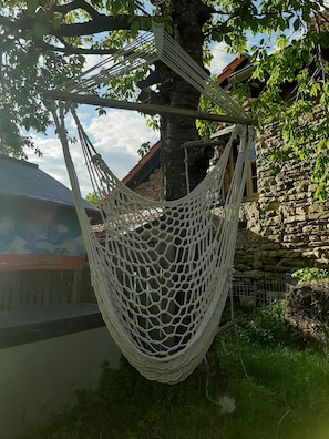 A hammock hanging between trees in a garden area with a stone wall and a building with a sloped roof.