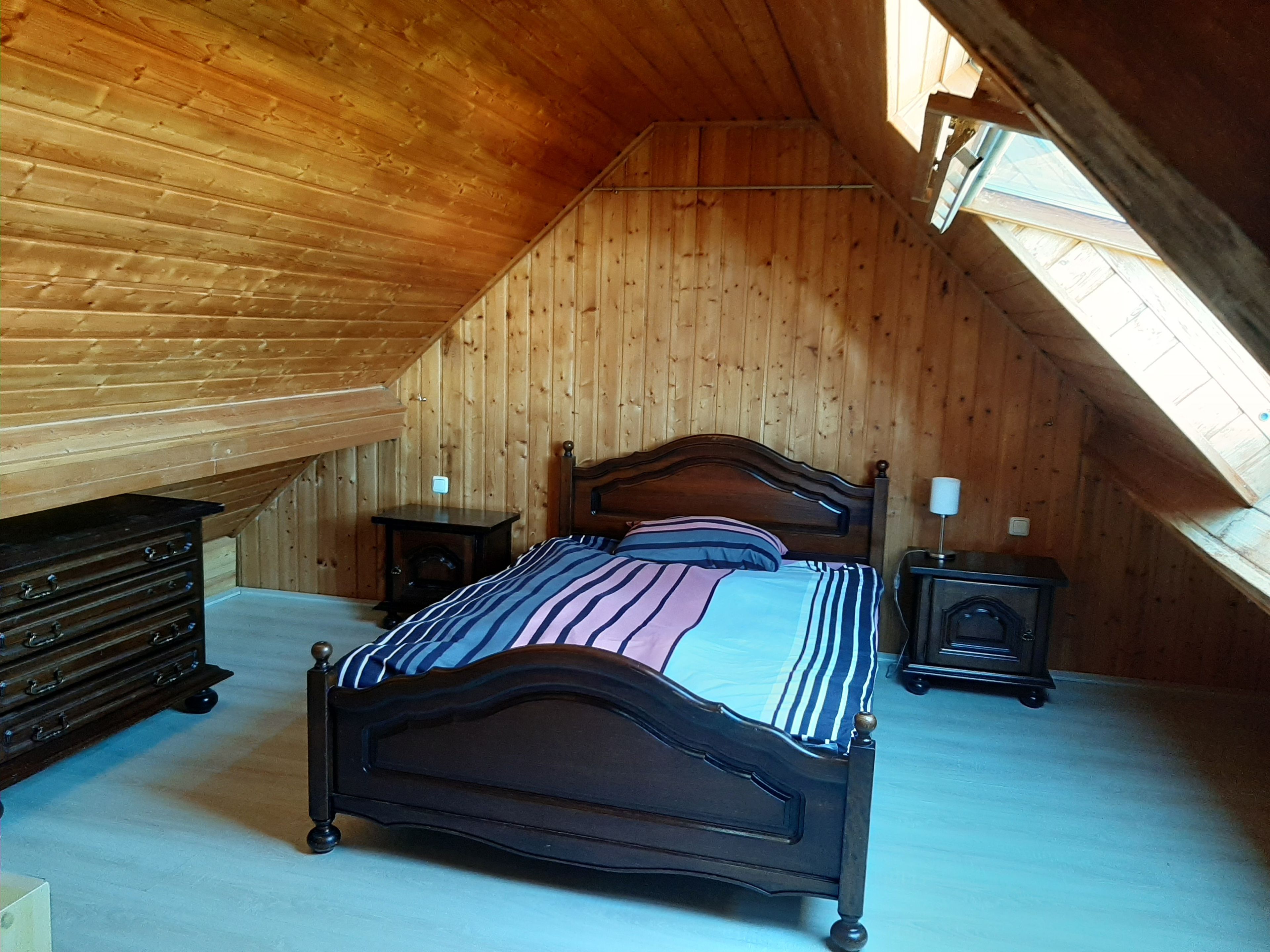 Ein Schlafzimmer mit schrägen Wänden, einem Bett mit Holz-Kopfteil, einer Kommode, einem Nachttisch und einer Lampe.