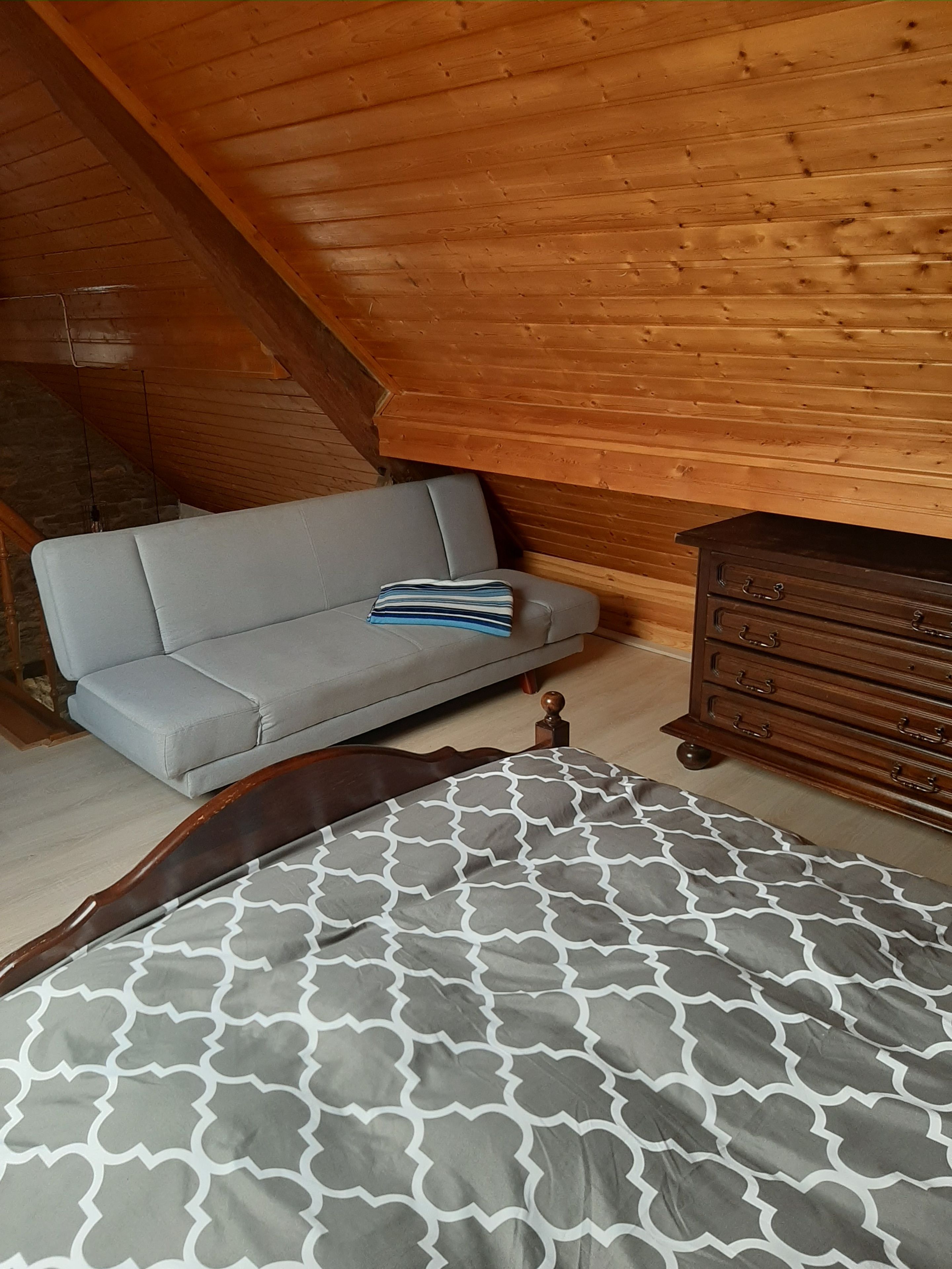 Ein Schlafzimmer im Loft-Stil mit schrägem Dach, einem grauen Sofa, einer Holzkommode und einer Betttdecke mit Muster.