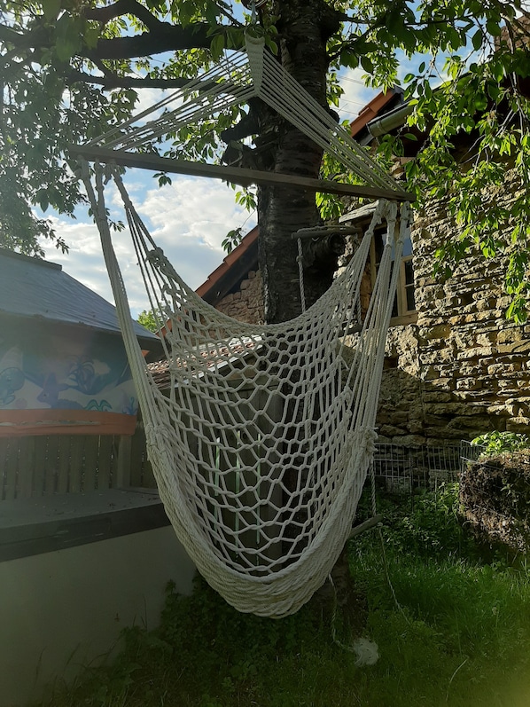 A hammock hanging between trees in a garden area with a stone wall and a building with a sloped roof.