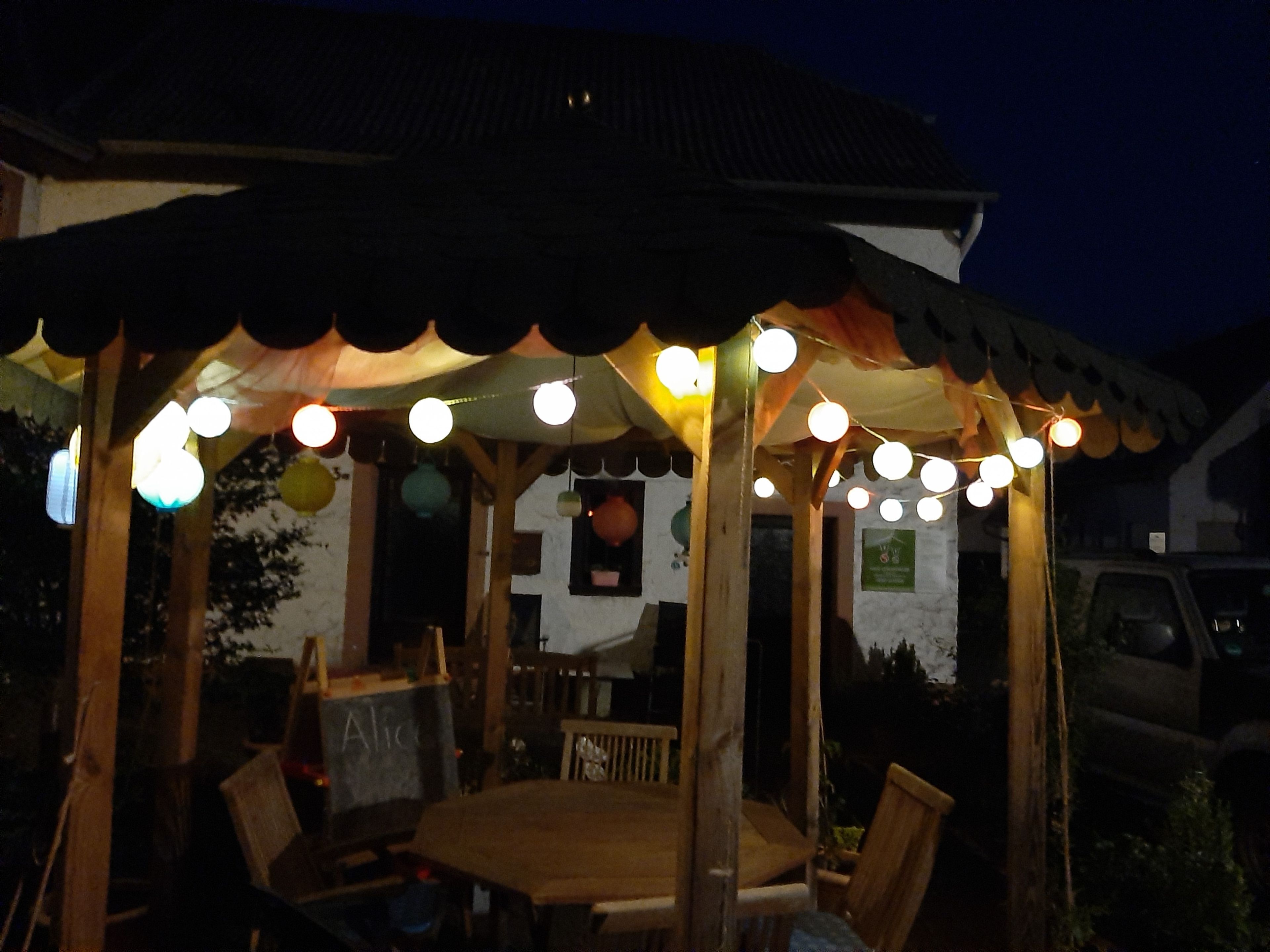 Ein hölzerner Pavillon mit einem Ziegeldach, umgeben von Stühlen und einem Tisch, mit Lichterketten beleuchtet.