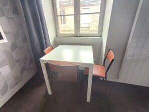 A small dining area with a table and two chairs, a window with a view of buildings, and a patterned wall. - Charmant studio aux Chartrons avec climatisation (Bordeaux)