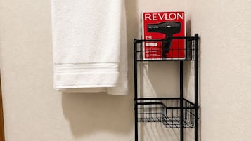 Ein Badezimmer mit einem Handtuch, das an einem schwarzen Handtuchhalter hängt, einer Revlon-Box auf einem schwarzen Metallregal und braunen Fliesen.
