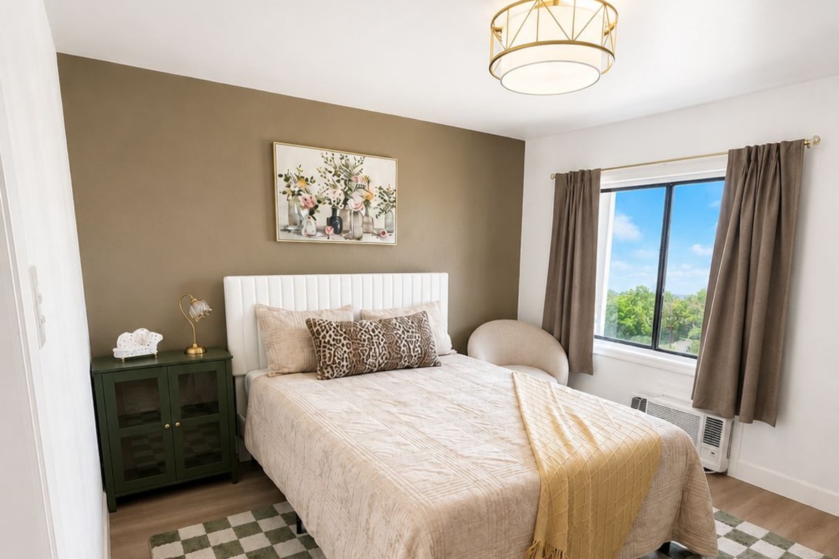 A bedroom with a bed, a nightstand, a lamp, a painting, a window with curtains, and a checkered rug.