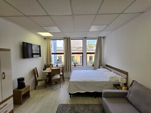 A hotel room with a bed, a desk, a chair, a sofa, a TV, and a window with curtains. - Fully Equipped Apartment in Central Area (Montevideo)