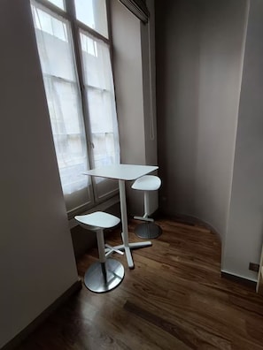 A corner room with a white table and two white bar stools, a window with sheer curtains, and wooden flooring. - Studio cosy quartier Chartrons (Bordeaux)