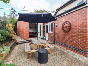 A patio area with a wooden table and benches, a black trash bin, and a brick wall with a clock. - The Snug (York)