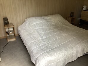 Ein ordentlich bezogenes Bett mit weißer Daunendecke, ein Nachttisch mit Lampe und eine kleine Ablagefläche mit Telefon und Ladegerät.