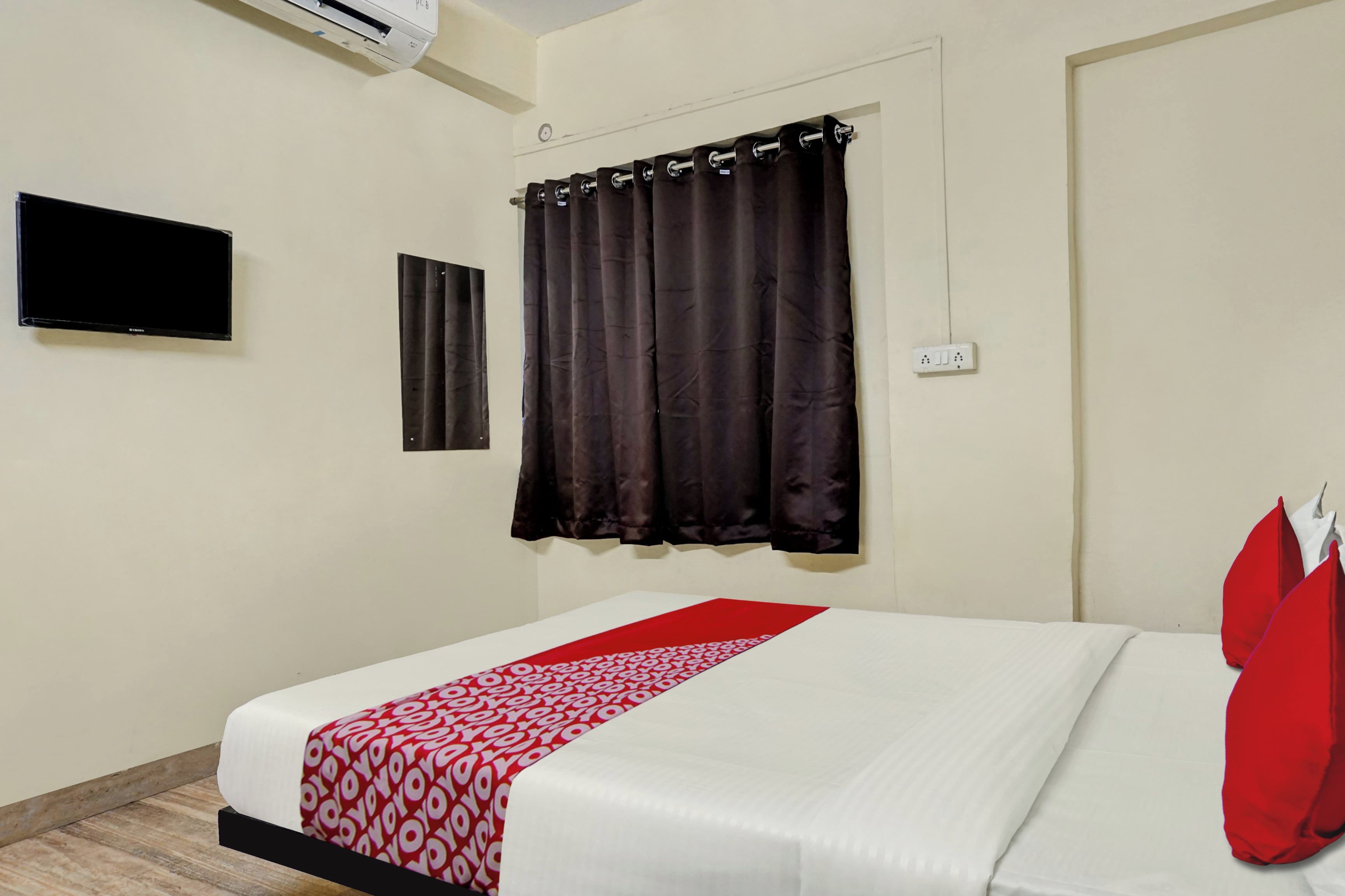 A hotel room with a bed, a flat-screen TV mounted on the wall, a window with dark curtains, and a power outlet.