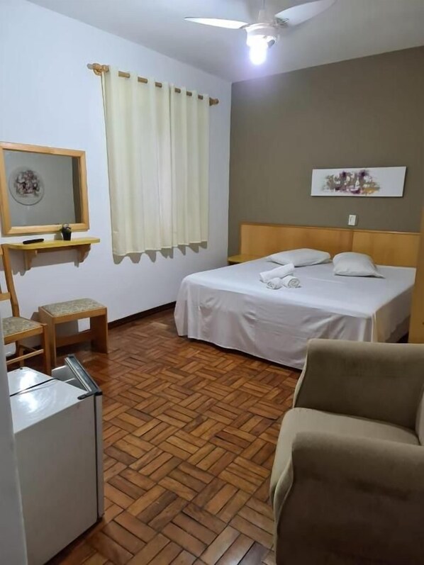 A hotel room with a bed, a chair, a small table, a mirror, a ceiling fan, and a window with curtains.