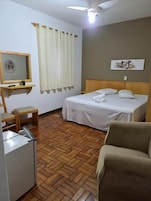 Habitación de hotel con cama, una silla, una mesita, un espejo, ventilador de techo y ventana con cortinas.