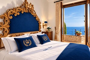 Ein Kingsize-Bett mit blauer Kopfteil und passenden Kissen, ein Nachttisch mit Lampe sowie Meerblick vom Balkon.