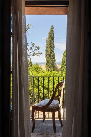 Ein Balkon mit einem Holzstuhl, Vorhängen und Blick auf eine grüne Landschaft mit Bäumen und Hügeln.