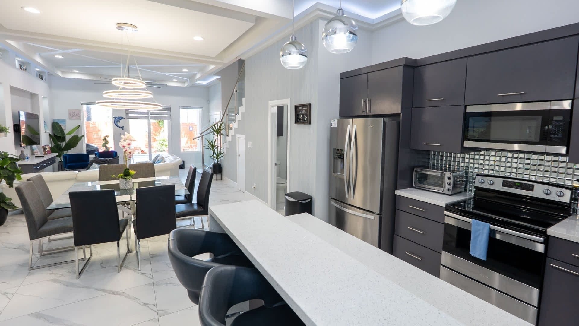 A modern kitchen with stainless steel appliances, a dining area with a glass table, and a living space with a sofa and plants.