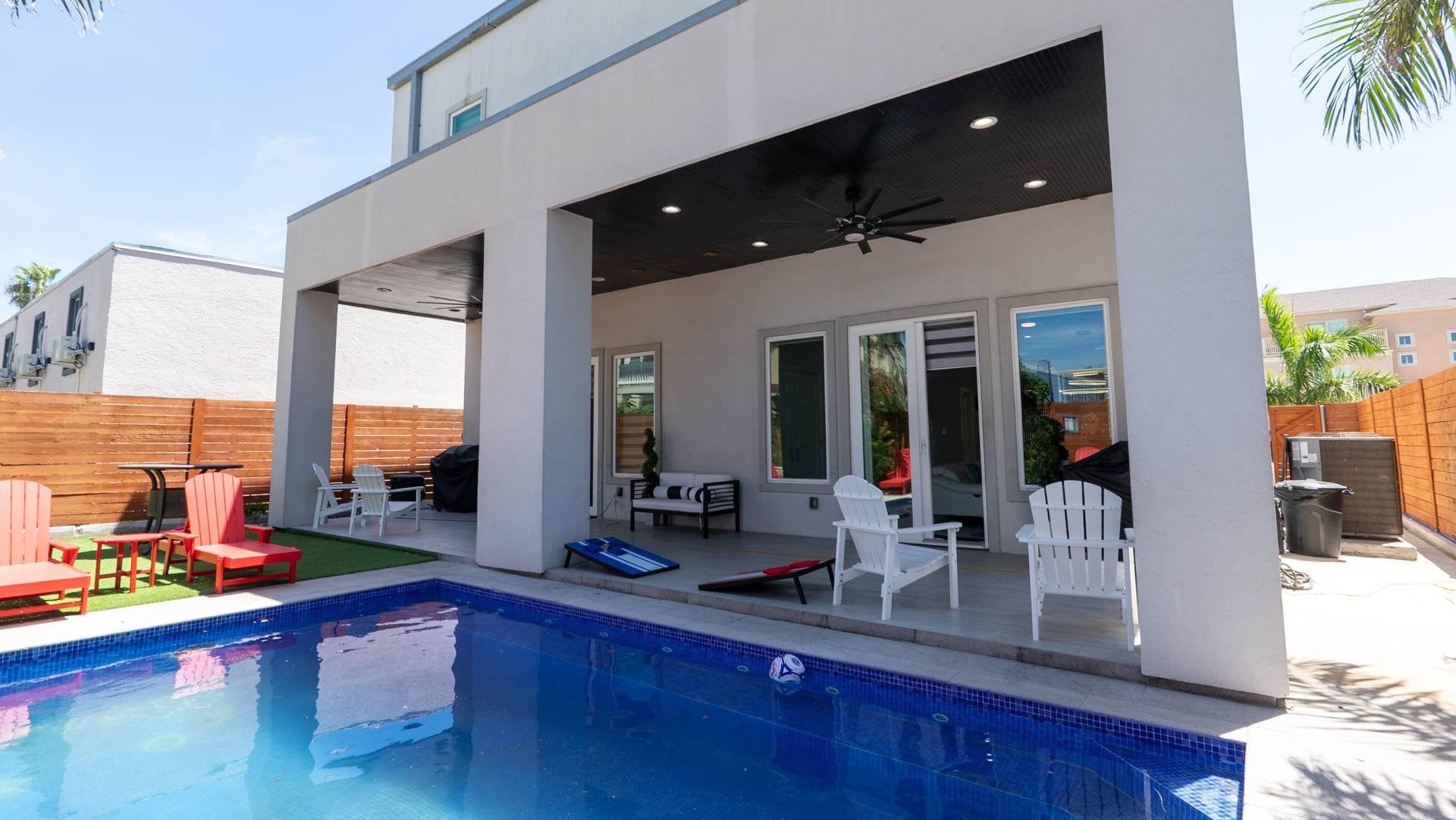 A modern outdoor pool area with a covered patio, seating, and a ceiling fan.