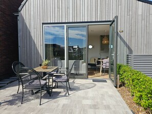 Eine Terrasse im Freien mit Tisch und Stühlen, eine Glastür, die zu einem Innenraum führt, und eine kleine Gartenanlage.