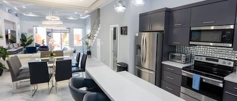 A modern kitchen with stainless steel appliances, a dining area with a glass table, and a living space with a sofa and plants.
