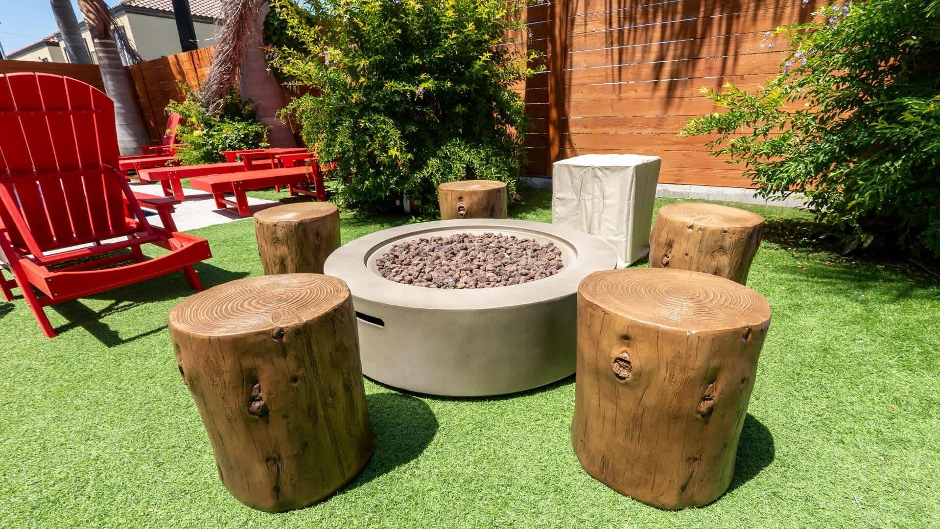 A fire pit surrounded by wooden stools and red chairs in a garden setting.