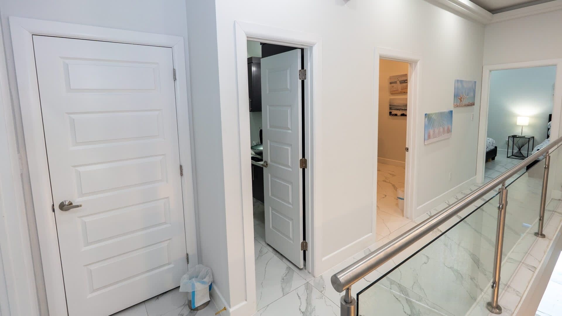 Een gang met een witte deur, een glazen balustrade en uitzicht op een slaapkamer met een bed en nachtkastje.