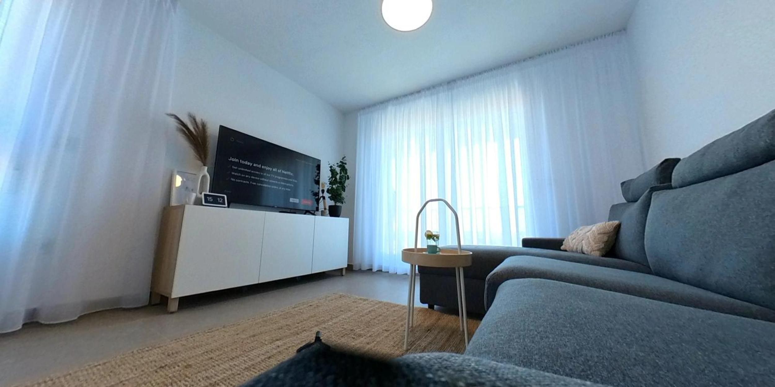 Una sala de estar moderna con un sofá gris, un televisor de pantalla plana sobre un mueble blanco y una pequeña mesita con una planta y una taza.