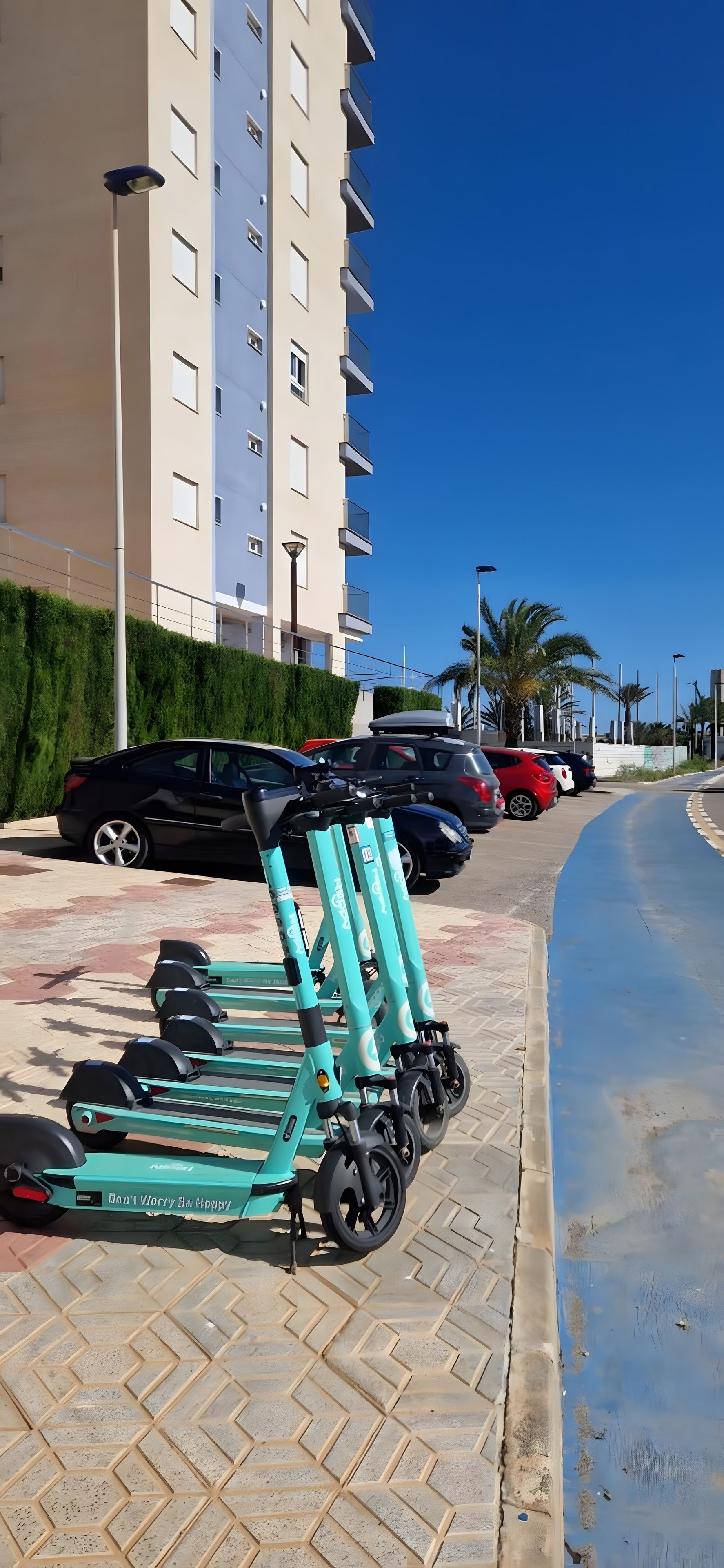 Un espace de stationnement avec plusieurs trottinettes électriques, une rangée de voitures garées et un immeuble doté de nombreux balcons.