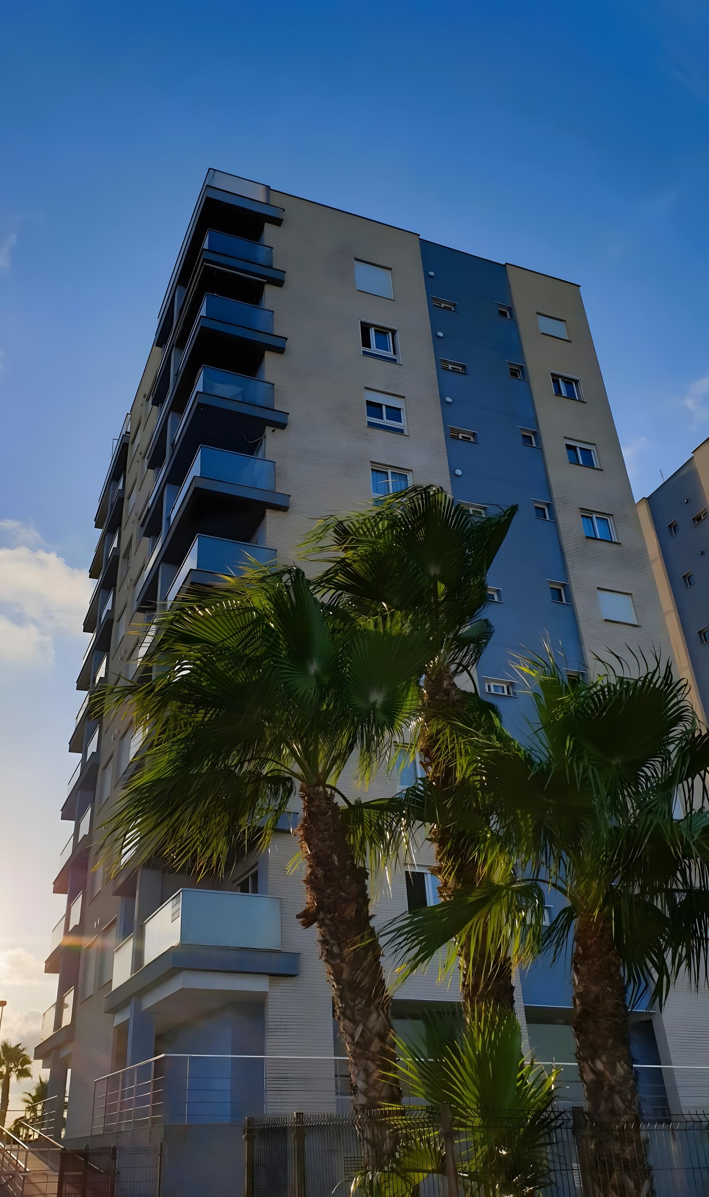 A multi-story building with balconies and windows, surrounded by palm trees.