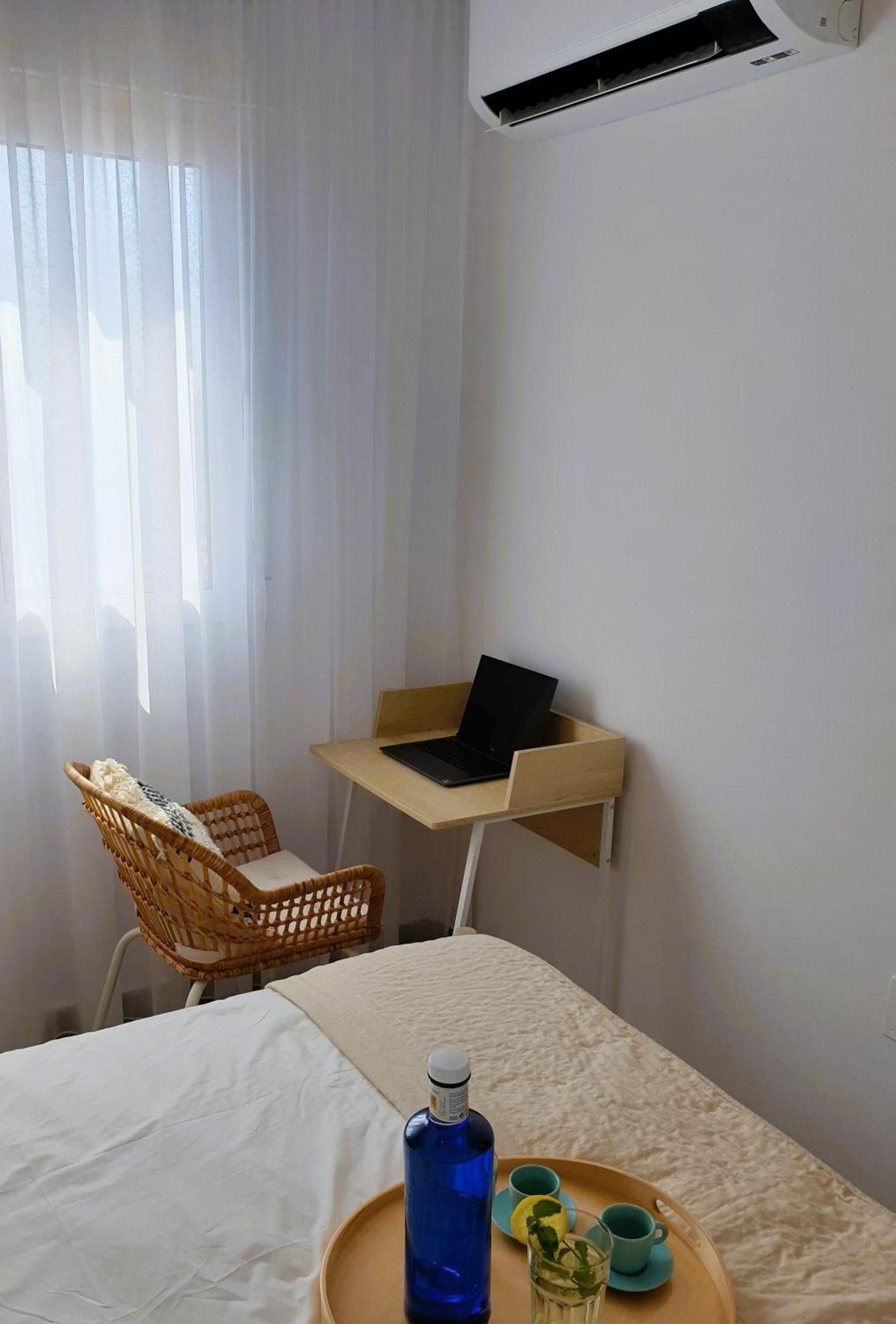 Une chambre d’hôtel avec un lit, un bureau avec un ordinateur portable, une chaise en osier et un plateau avec une bouteille et des verres.
