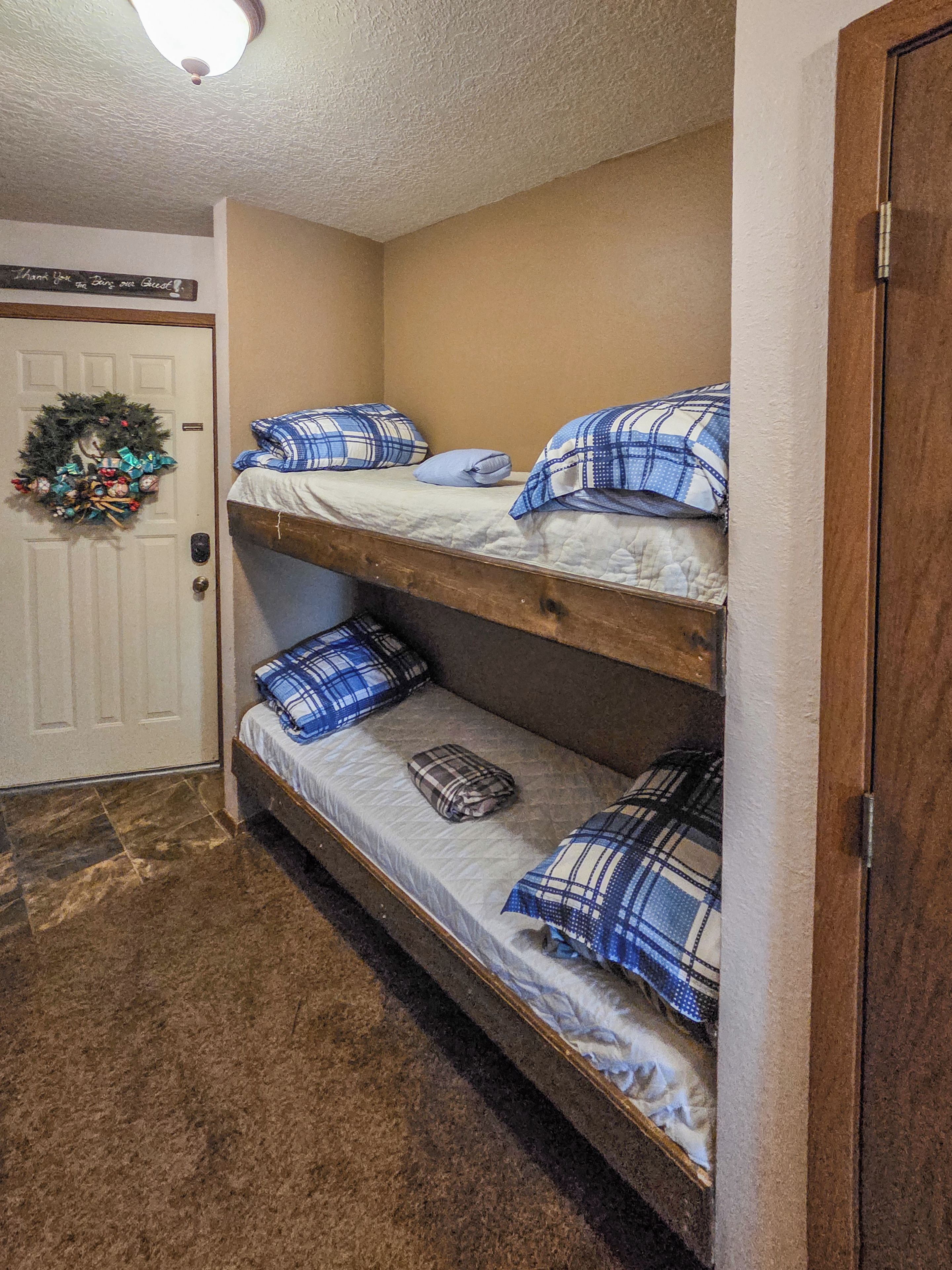A bunk bed with blue and white bedding, a door with a wreath, and a carpeted floor.