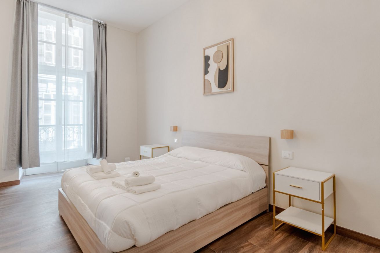 A modern hotel room with a large bed, bedside tables, a window with curtains, and a wall-mounted artwork.