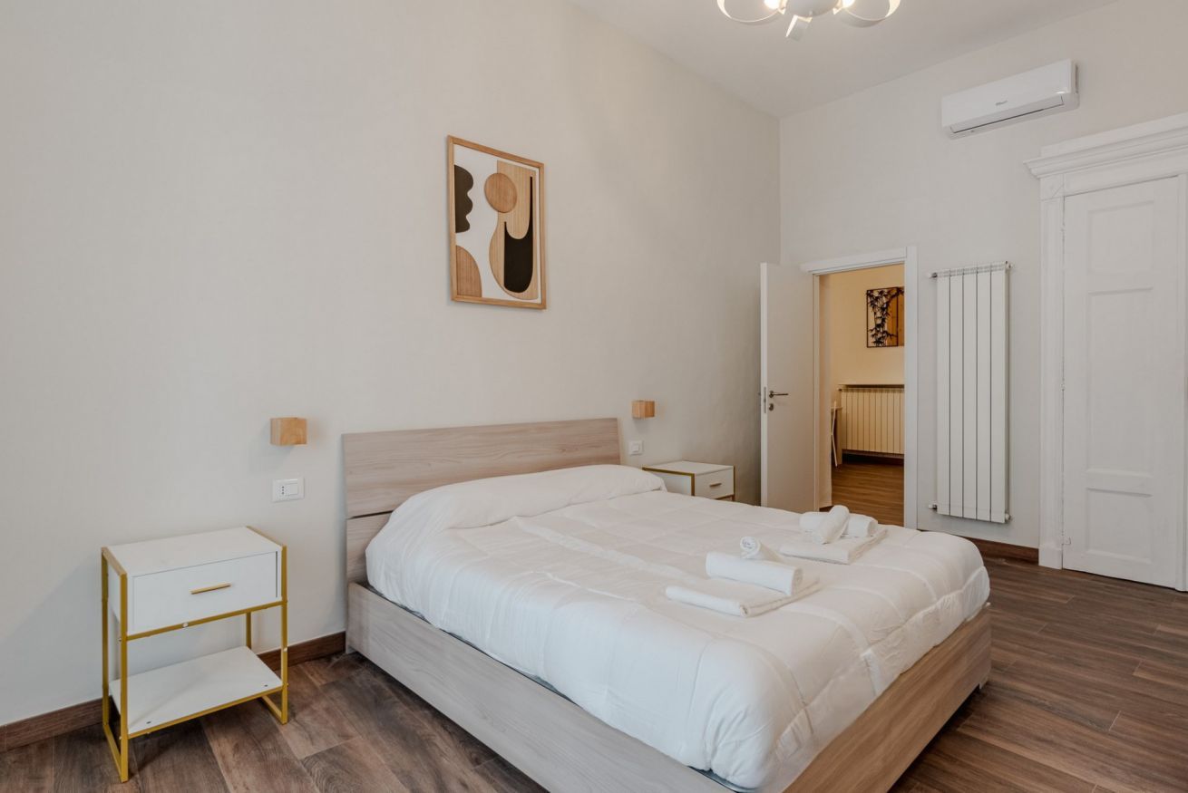 Habitación de hotel moderna con una cama grande, una mesita de noche, una obra de arte colgada en la pared y una puerta abierta que da a otra habitación.