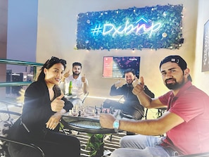 A group of people sitting around a table with a chessboard, a neon sign, and a screen displaying a menu. - Pathfinder Partition Room in Tecom (Dubai)