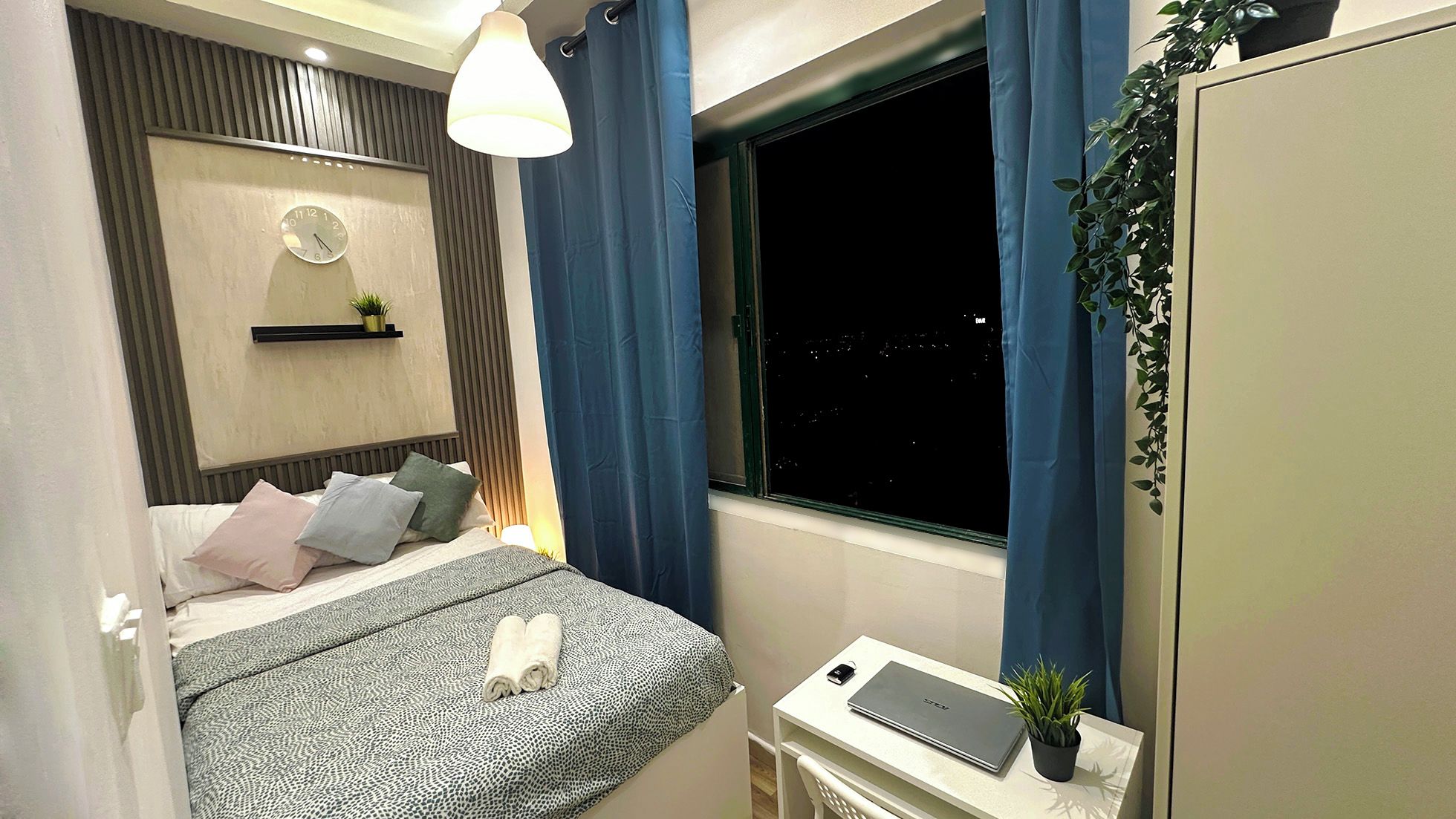 A bedroom with a bed, a bedside table with a laptop, a window with curtains, a wall clock, and a small plant.