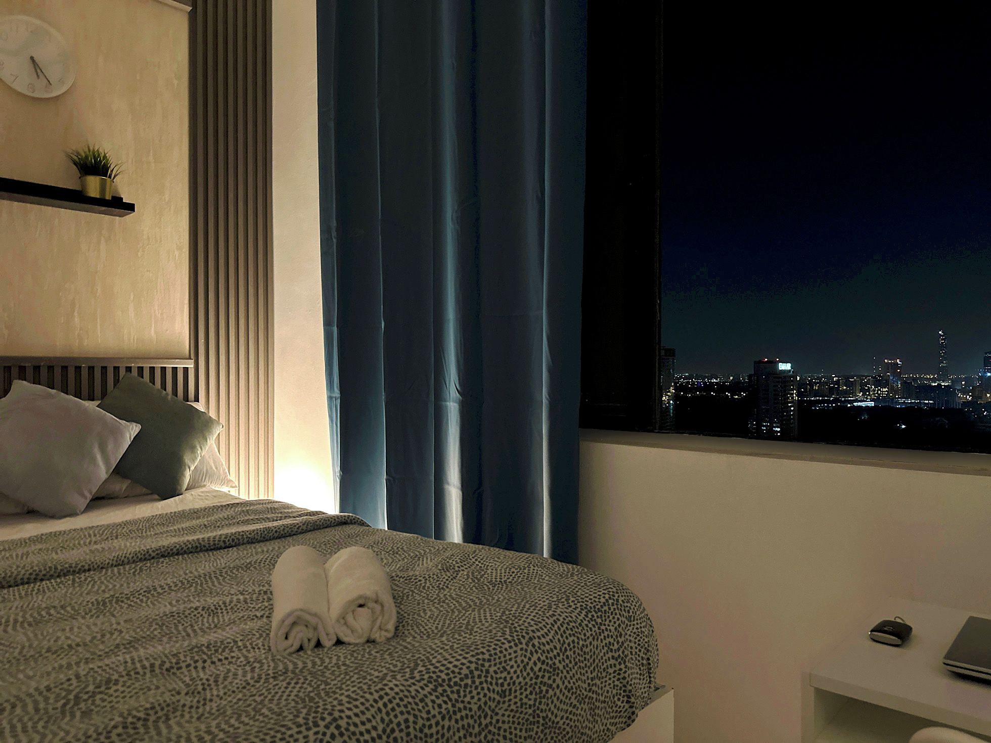Een hotelkamer met een bed, nachtkastje, raam met uitzicht over de stad en een klok aan de muur.